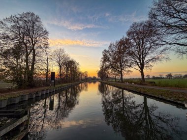 Rijkevorsel, Antwerpse Kempen, Belçika, günbatımı kanalı aynalı su, ağaçlı kıyılar ve uzak kilit çerçeveli altın saat gökyüzü, sakin yansıtıcı yüzey