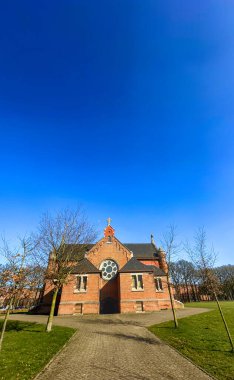 Merksplas, Antwerp, Belçika, 18 Nisan 2025, göz kamaştırıcı ve büyüleyici tuğla bir kilise canlı mavi gökyüzünün altında güzel ve gür ağaçlarla çevrili duruyor.