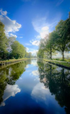 Huzurlu ve sakin bir nehir manzarası sergilenen canlı yeşillik çarpıcı bulut yansımalarıyla çelişiyor.