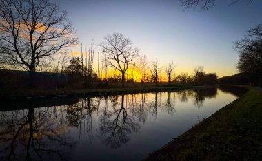 Gün batımında büyüleyici ve sakin bir nehir manzarası. Ağaçların ve canlı gökyüzünün güzel yansımaları.