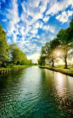 Gün batımında çarpıcı, bulutlu bir gökyüzünün yanında canlı yeşillikler barındıran güzel, sakin bir nehir manzarası.