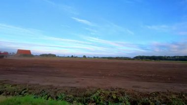 Rijkevorsel, Antwerpse Kempen, Belçika, gündoğumu tarlası traktör sürüşü, ufukta kırmızı çiftlik evi, açık kahverengi toprak açık mavi gökyüzü altında, çizgili bulutlar, yumuşak sabah