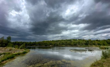 Derin derin düşünen fırtına bulutları sessiz gölün üzerinde toplanıyor, karanlık dramatik gökyüzü suya yansıyor, engebeli kıyı şeridi ve uzak ağaç hattı