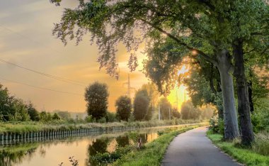 Gündoğumu kanalı yolu ağaçların arasından süzülen sıcak ışıkla sabah koşucularını davet ediyor, çiğ taneli otlar, sakin yansımalar.