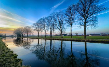 Huzurlu ve sakin bir nehir manzarası. Siluetli ağaçlar gün batımında sakin suları incelikle yansıtıyor.