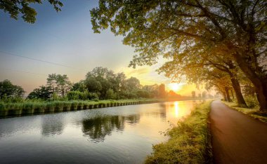Gün batımında sakin ve sakin bir nehir manzarası, canlı, renkli ağaçlar ve huzurlu yürüyüş yolları ile çevrili.