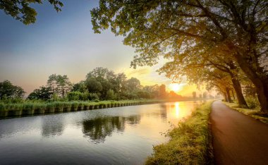Güzel sakin bir günbatımı sahnesi sakin, dingin bir nehri yansıtıyor, yemyeşil, canlı yeşilliklerle çevrili.