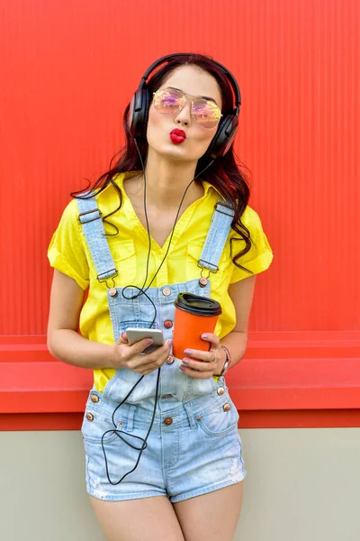 Beautiful smiling woman with headphones listens to music over red background.  Fashion woman in sunglasses outdoor.