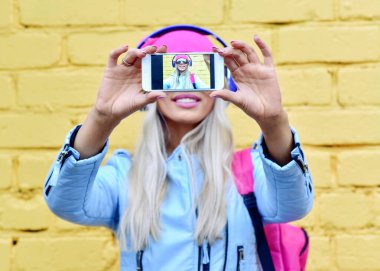 Moda kadını, şehrin renkli sarı arka planında akıllı telefon ekranında kendi portresini yapıyor.