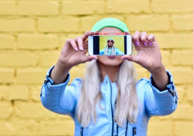 Moda kadını, şehrin renkli sarı arka planında akıllı telefon ekranında kendi portresini yapıyor.