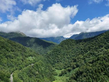 A mountain range with a forest in the foreground and a road in the background. The sky is cloudy, but the sun is still shining through. Scene is peaceful and serene, with the lush green trees