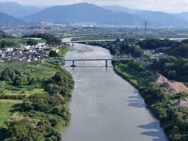 A beautiful river with a bridge spanning across it, surrounded by a lush green landscape. In the background, there is a city with tall buildings and a bustling atmosphere