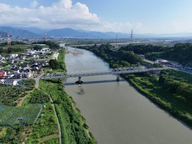 Bir köprü, arka planında yeşil ve kahverengi bir manzara olan bir nehri kapsar. Köprü evler ve ağaçlarla çevrili.