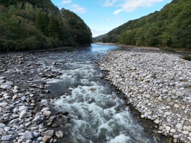 Kayalık bir kıyı şeridi ve açık mavi bir gökyüzü olan bir nehir. Su hızlı akıyor ve kayalar kıyı boyunca yayılıyor.