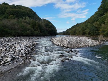 Kayalık bir kıyı şeridi ve açık mavi bir gökyüzü olan bir nehir. Su akıyor ve kayalar kıyıya dağılıyor.