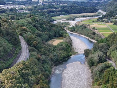 A river runs through a forest and a road runs alongside it. The road is a winding road that is surrounded by trees