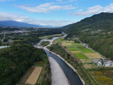 Bir nehir, sağ tarafında bir kasaba olan bir vadiden geçer. Gökyüzü mavi ve dağlar arka planda.