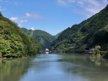 A river with a green forest on either side. The water is calm and clear. The sky is blue with some clouds