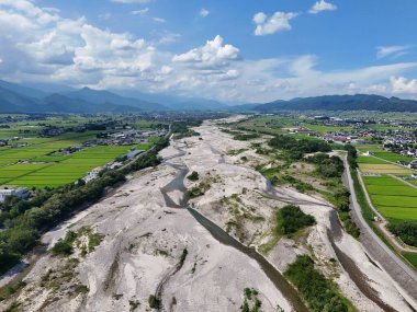 Kırsal bir alandan geçen büyük, kumlu bir nehir. Gökyüzü çoğunlukla açık ve birkaç bulut etrafa saçılmış durumda. Manzara çoğunlukla düzdür, bazı tepeler uzaktadır.