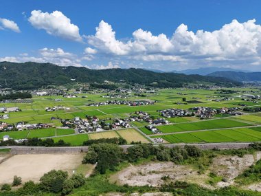Yeşil tarlaları ve evleri olan bir kırsal bölge. Gökyüzü birkaç bulutla daha açık.