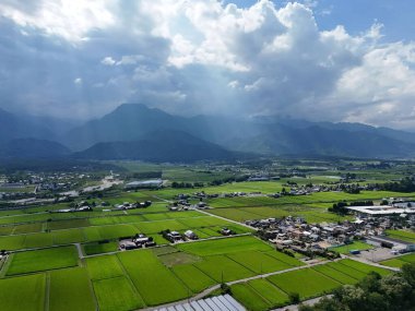 A large field of green grass with a few houses in the distance. The sky is cloudy and the sun is shining through the clouds