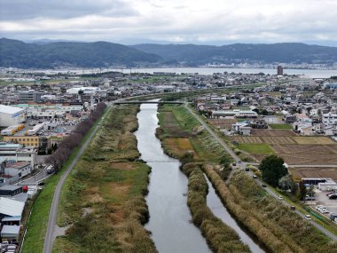 Her iki tarafında evler ve binalar olan bir şehrin içinden bir nehir akıyor. Nehir çimenler ve ağaçlarla çevrili ve yol boyunca giden arabalar ve kamyonlar var. Huzurlu bir sahne.