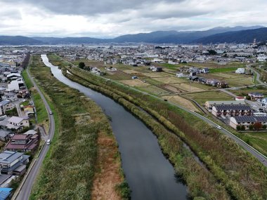 Her iki tarafında evler ve binalar olan bir kasabanın içinden bir nehir akıyor. Nehir çimenler ve ağaçlarla çevrili ve yol boyunca giden arabalar ve kamyonlar var. Huzurlu bir sahne.