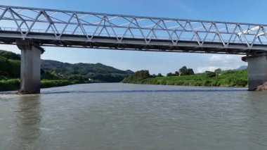 Metal köprünün altından akan nehir yeşilliklerle çevrili.