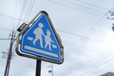 Beyaz üçgenli mavi bir tabela ve caddede yürüyen iki çocuk. Tabela bir direğin üzerinde ve kablolarla çevrili.