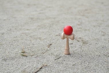 Kumlu bir plajda tahta bir oyuncak oturuyor. Oyuncak beyaz merkezli kırmızı bir top. Sahne huzurlu ve sakin, oyuncak kumlu arka planda duruyor.