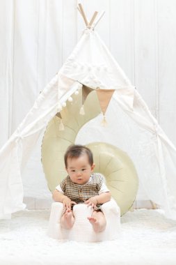 Çadırın önündeki taburede bir bebek oturuyor. Çadır bayraklar ve büyük bir balonla süslenmiştir. Bebek ekose bir gömlek giyiyor ve kameraya bakıyor. Sahne çok eğlenceli.