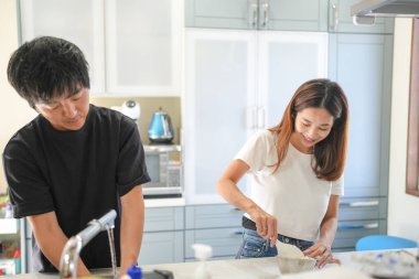 Bir adam ve bir kadın mutfaktalar, erkek bir şeyler kesiyor ve kadın gülümsüyor.