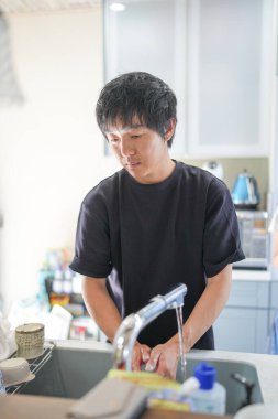 Bir adam lavaboda ellerini yıkıyor. Lavabo bulaşık ve şişelerle dolu. Adam siyah bir gömlek giyiyor.