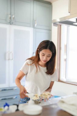 Bir kadın mutfakta tatlı pişiriyor. O beyaz bir gömlek ve kot pantolon giyiyor. Mutfakta buzdolabı, fırın ve lavabo var.
