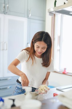 Bir kadın mutfakta, gülümsüyor ve yemek hazırlıyor. Beyaz bir gömlek ve kot pantolon giyiyor.