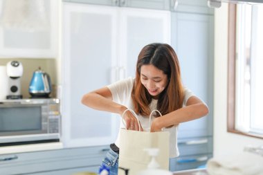 Bir kadın mutfakta bir çanta açıyor. Gülümsüyor ve mutlu. Mutfak mikrodalga, tost makinesi ve fırın gibi aletlerle donatılmıştır.