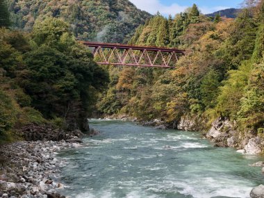 Bir köprü yeşil ve mavi bir su yolu ile bir nehri kapsar. Köprü kırmızıdır ve nehrin tüm genişliğini kapsar. Köprü ağaçlar ve kayalarla çevrili.