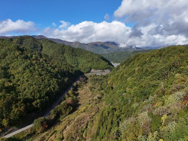 İçinden yol geçen bir dağ sırası. Yol ağaçlarla çevrili ve gökyüzü bulutlu.