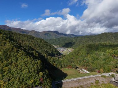 Önünde orman olan bir dağ sırası. Gökyüzü bulutlu ve güneş bulutların arasında parlıyor.