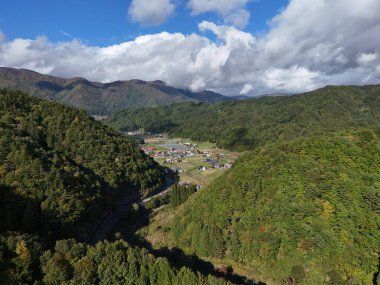Aralarında vadi olan bir dağ sırası. Vadi evler ve ağaçlarla dolu. Gökyüzü bulutlu ve güneş parlıyor