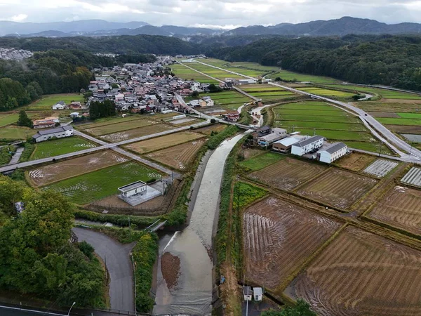 Kırsal bir bölge ve içinden bir nehir geçiyor. Evler küçük ve yayılmış. Gökyüzü bulutlu.