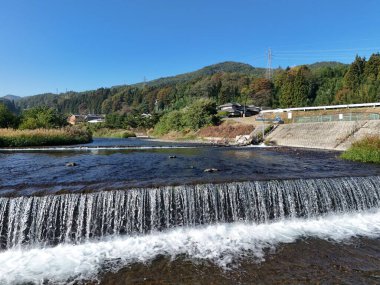 Temiz bir su akıntısı olan güzel bir şelale. Su ağaçlarla ve arka planda küçük bir kasabayla çevrilidir. Huzurlu ve huzurlu bir sahne.