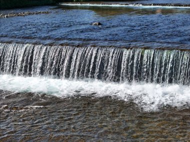 Bir şelale nehre akıyor. Su temiz ve kayalar sahnenin her tarafına dağılmış.