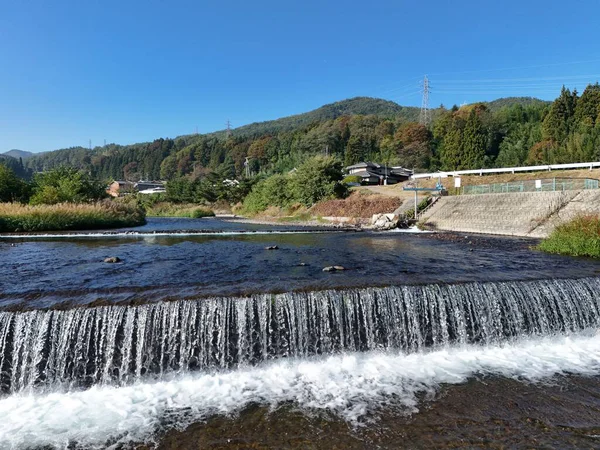 Temiz bir su akıntısı olan güzel bir şelale. Su ağaçlarla ve arka planda küçük bir kasabayla çevrilidir. Huzurlu ve huzurlu bir sahne.