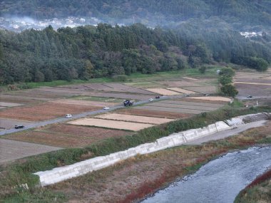 Kırsal bir bölge ve içinden bir nehir geçiyor. Nehir bir toprak yol ve bir çitle çevrilidir. Yol boyunca park edilmiş birkaç araba var.