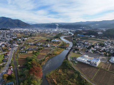 İçinden nehir akan bir şehir. Nehir çiftlik arazileri ve evlerle çevrilidir. Gökyüzü açık ve güneş parlıyor.