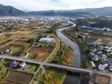Kırsal bir bölge ve içinden bir nehir geçiyor. Nehir çiftlik arazileri ve evlerle çevrilidir. Gökyüzü açık ve güneş parlıyor.