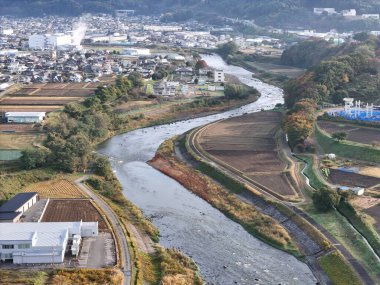Arka planda evler ve binalar olan bir kasabanın içinden bir nehir akıyor. Nehir kahverengi ve bulanık. Yolun kenarında park etmiş birkaç kamyon var.