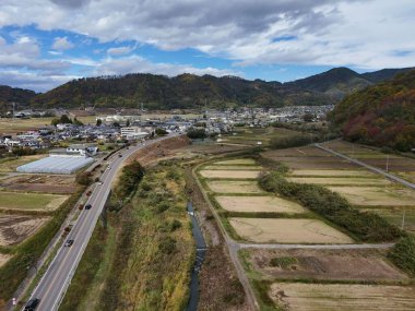 Bir yol ve birkaç evin olduğu kırsal bir bölge. Gökyüzü bulutlu ve güneş parlıyor