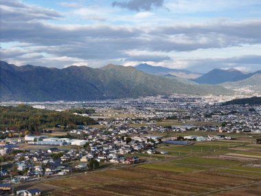 Arkasında dağ olan bir şehir. Şehir evler ve binalarla dolu. Gökyüzü bulutlu ve güneş görünmüyor.